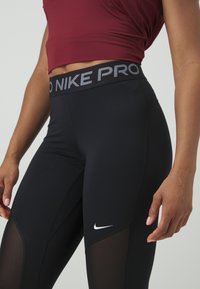 Mustat Nike Pro leggingsit, joissa on korkea vyötärö harmaalla tekstillä, verkkopaneelit sivuilla ja pieni valkoinen logo helman lähellä.