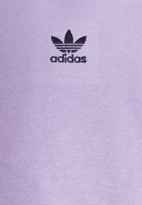 Lila Baumwoll-T-Shirt mit schwarzem besticktem Adidas-Logo und drei Blattakzenten im linken Brustbereich. Glatte Textur, lässiges Design.