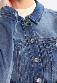 Chaqueta de mezclilla en azul claro con cierre de botones, cuello clásico y dos bolsillos en el pecho. Presenta costuras visibles y botones de metal.