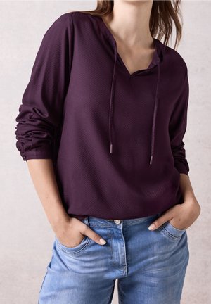 Kvinde iført en lilla, tekstureret bluse med lange ærmer med snore og lyseblå jeans, hænderne stukket i frontlommerne.