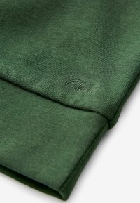 Tessuto di felpa verde scuro con una texture liscia. Presenta un polsino ripiegato e un nome del marchio ricamato in una tonalità sobria sulla manica.