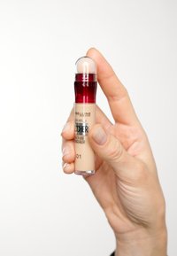 Correcteur fluide beige dans un tube en plastique transparent, avec un capuchon rouge et un applicateur en mousse. L'étiquette affiche la marque et le nom du produit.