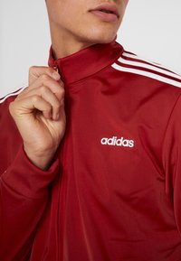 Chaqueta de entrenamiento roja de Adidas con rayas blancas en los hombros, cuello con cremallera y logo situado en el lado izquierdo. Tejido suave y ligero.