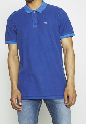 Mężczyzna w niebieskiej koszulce polo z małym logo w kolorach czerwonym, białym i niebieskim na piersi oraz jasnoniebieskich dżinsach.