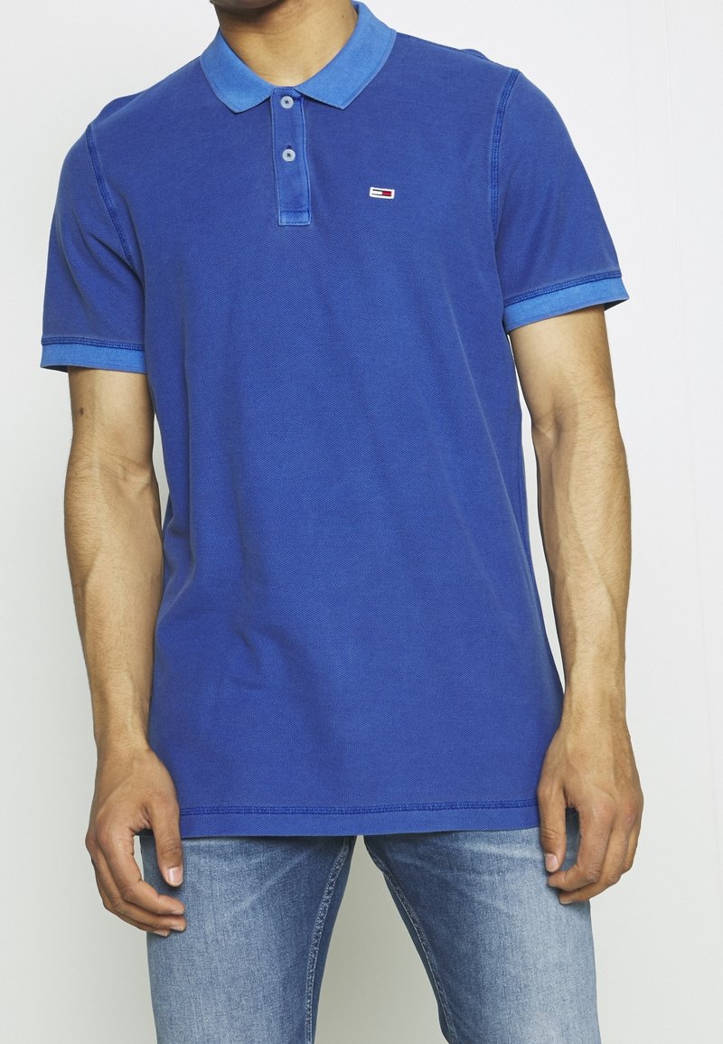 Homme portant un polo bleu avec un petit logo rouge, blanc et bleu sur la poitrine et un jean bleu clair.