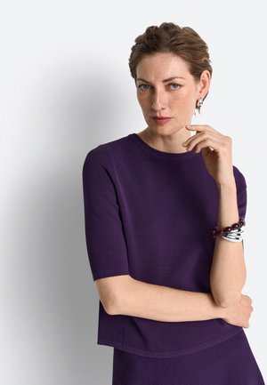 Kvinde med kort hår iført en lilla top og matchende nederdel, som rører ved sit ansigt med den ene hånd og bærer sølv- og mørkerøde armbånd.