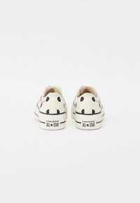 Bijele platnene tenisice s crnim točkicama, ravnim vezicama i gumiranim potplatom. Imaju otisnuti crveni logo sa strane i "Converse All Star" oznaku na peti.