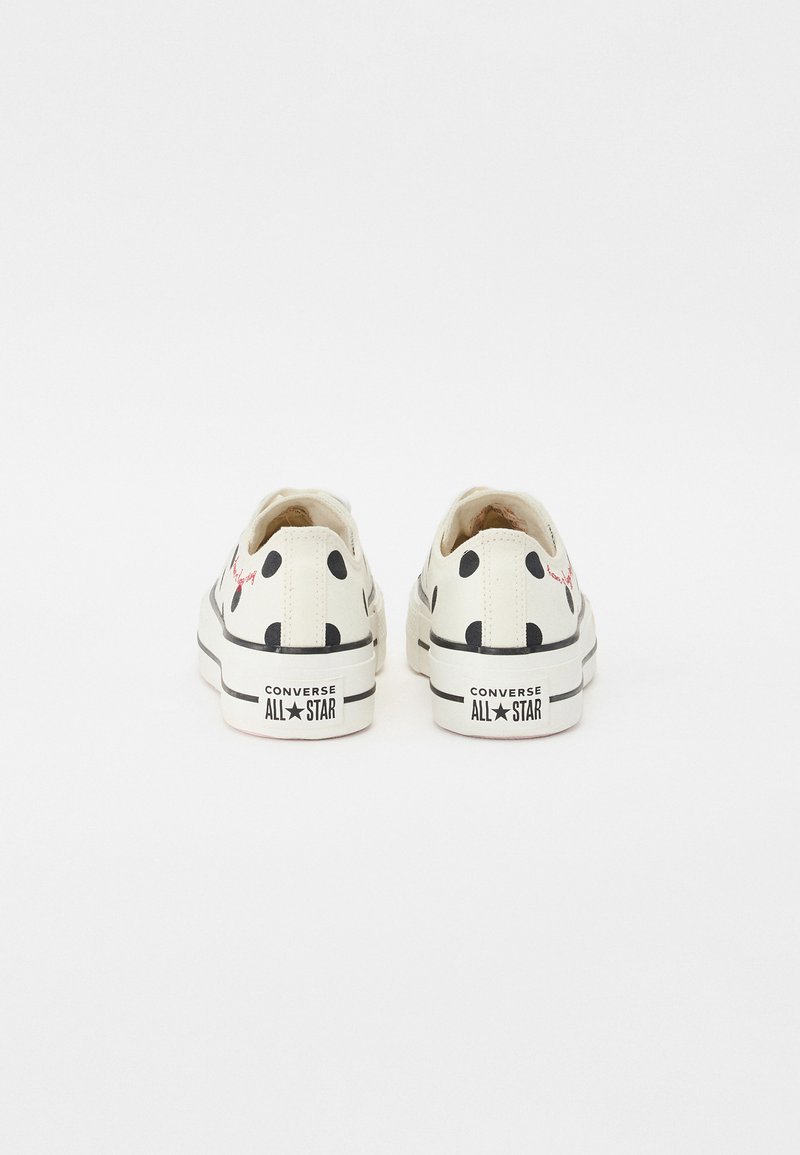 Zapatillas de lona blancas con puntos negros, cordones planos y suela de goma. Presentan un logo rojo impreso en el lateral y la marca "Converse All Star" en el talón.