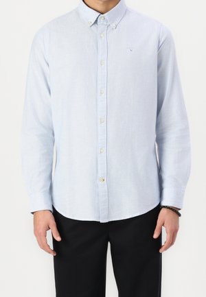 Homme portant une chemise bleu clair à manches longues boutonnée et un pantalon noir, debout, les mains détendues le long du corps.