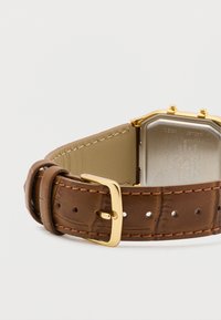 Brązowy skórzany pasek do zegarka z złotą metalową klamrą, z ośmiokątną srebrną tarczą zegarka z wygrawerowanymi detalami i brązowymi akcentami.