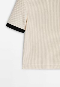 Haut tricoté à manches courtes de couleur crème avec un motif gaufré texturé et une bande noire contrastante sur le bord de la manche.