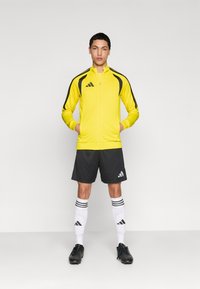 Κίτρινο αθλητικό μπουφάν με φερμουάρ και μαύρες λεπτομέρειες, μαύρο σορτς και λευκές κάλτσες με ρίγες. Το μοντέλο φοράει μαύρα ποδοσφαιρικά παπούτσια και έχει τα χέρια στις τσέπες.