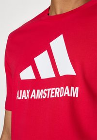 Rød bomulds T-shirt med et stort hvidt Adidas-logo med tre diagonale striber og fed tekst "AJAX AMSTERDAM" nedenunder logoet.