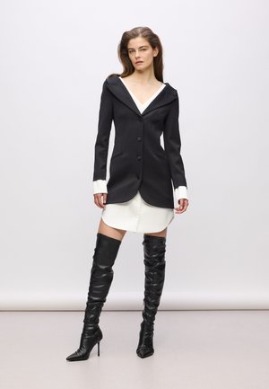 Black tailored blazer with a wide collar and three buttons, layered over a white shirt dress, paired with knee-high black leather boots.