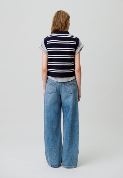 Maglione a righe navy con maniche corte grigie, abbinato a jeans in denim blu a gamba larga con dettagli ricamati sulle tasche posteriori.