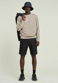 Felpa beige, pantaloni corti neri e un cappello a secchiello in denim. Modello che tiene una giacca, indossa scarpe da ginnastica nere e calze. Design semplice e casual.