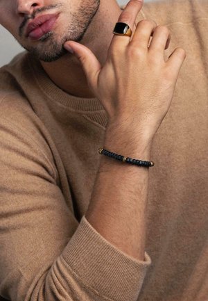 Beige sweater, dark beaded bracelet with gold accents on a wrist, and a large black ring on a finger. Skin tone is medium.