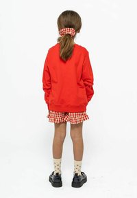 Červená mikina, kostičkovaná sukně s volánky, krémové ponožky se vzorem srdcí a černé boty. Vlasy stažené vzadu do gumičky v odpovídající barvě.