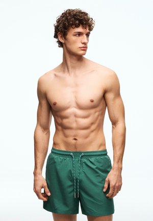 Młody mężczyzna z kręconymi włosami, ubrany w zielone kąpielówki, stoi bez koszulki i spogląda na bok na jednolitym jasnym tle.