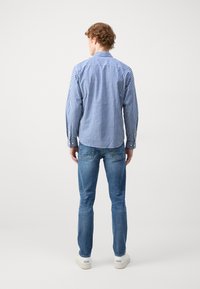 Blå rutete skjorte med lange ermer og krage, kombinert med lys blå jeans og hvite joggesko, sett fra baksiden mot en nøytral bakgrunn.