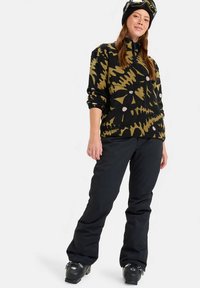 Νέος σε μπότες snowboard, με μαύρο παντελόνι και σχεδιασμένο παλτό σε μαύρο και ελιά, φοράει κράνος με γυαλιά.