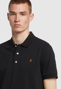 Zwarte polo met een gestructureerde afwerking, voorzien van een drie-knops sluiting en een klein oranje logo op de borst. Met kraag en korte mouwen.