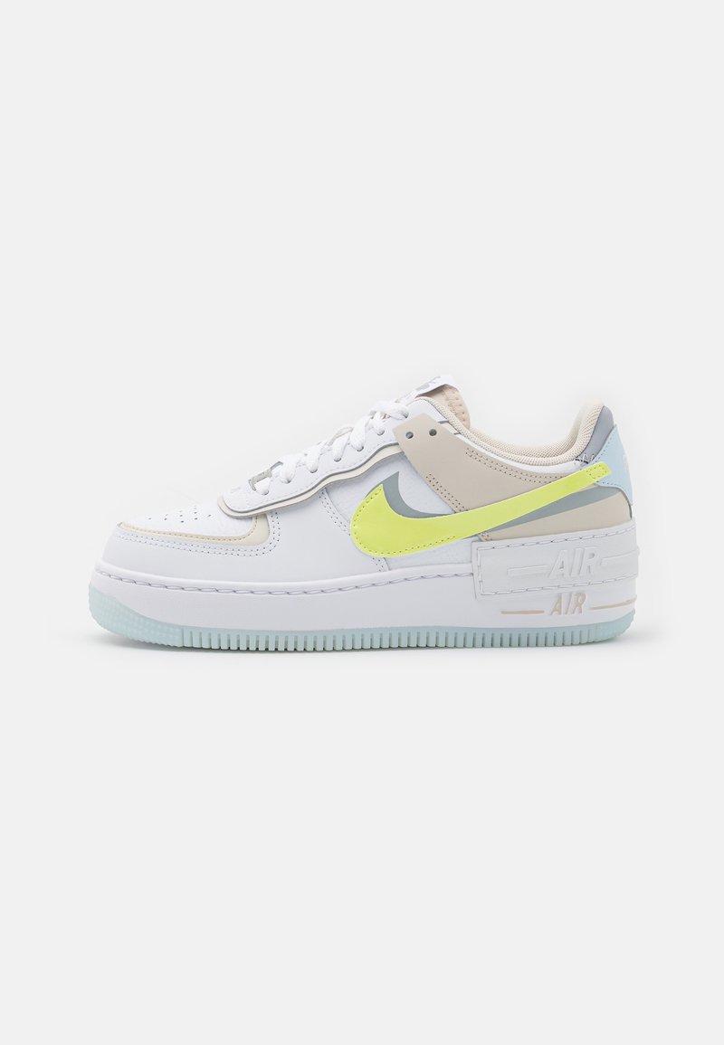Baskets blanches à tige basse avec des accents beiges, bleu clair et jaune fluo, dotées d'une semelle épaisse et d'un Swoosh Nike bien visible sur le côté.