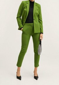 Conjunto de traje verde que incluye un blazer ajustado y pantalones entallados, confeccionados en una tela suave. Combinado con una blusa negra y un bolso de mano texturizado.