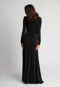 Kobieta o falujących brązowych włosach ubrana jest w długą, czarną dopasowaną sukienkę z marszczonymi rękawami, stojąc tyłem na tle jednolitego, jasnego tła.