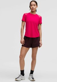 Ružový atletický tričko s krátkym rukávom a priesvitnými sieťovými detailmi, spárované s tmavohnedými šortkami. Model má na sebe biele a čierne atletické topánky.