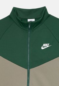 Žalia ir kreminė užtraukiama striukė su aukštu apykakle, turi Nike logotipą ir tekstūruotą audinį, demonstruojanti spalvų blokų dizainą.