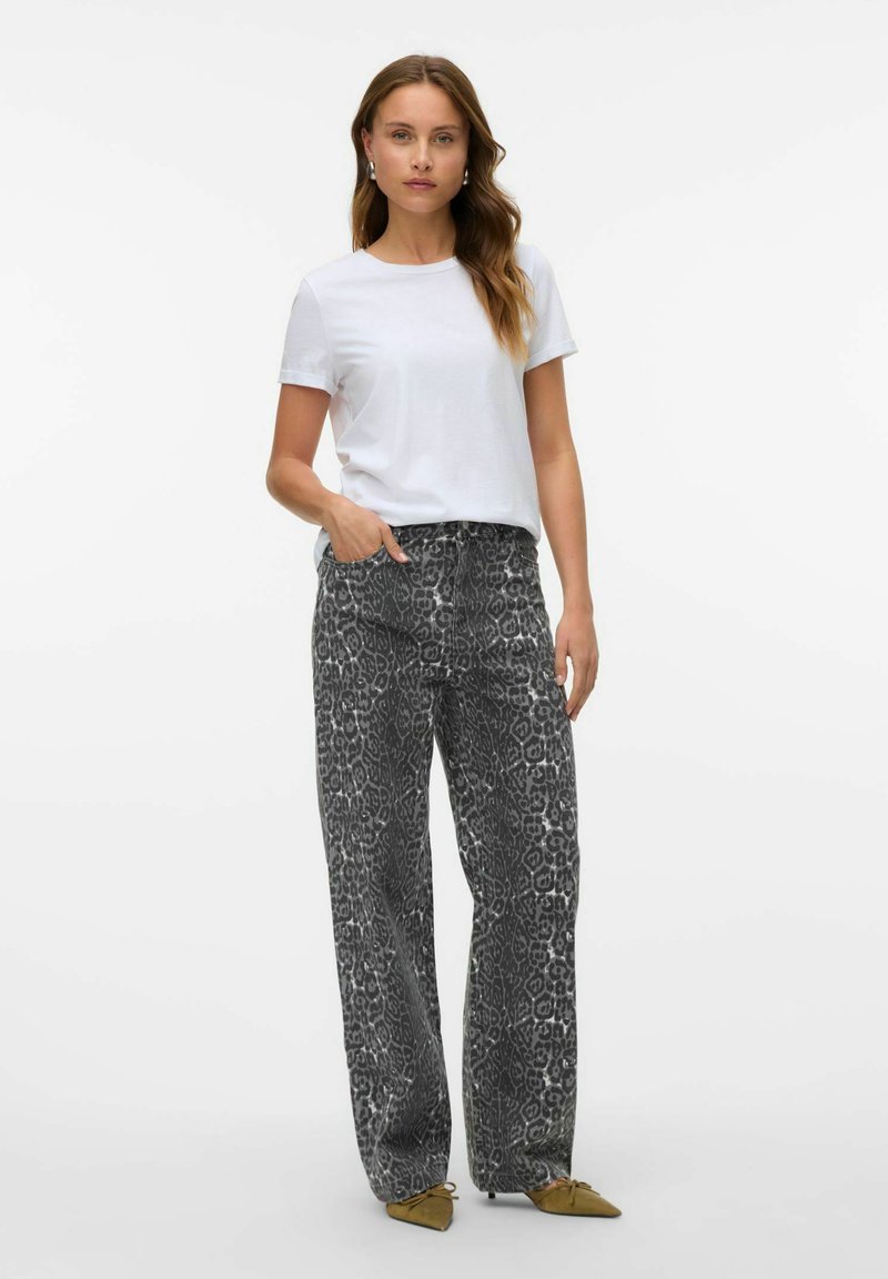 Femme portant un pantalon argenté imprimé léopard à jambes larges, un t-shirt blanc à manches courtes et des ballerines pointues moutarde, debout avec une main dans la poche.