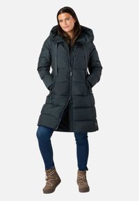 Sort pufferjakke med hætte, lynlås, to forlommer og knælængde. Båret med blå jeans og brune støvler.