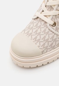 Gros plan sur la pointe d'une basket beige en tissu texturé, avec une semelle en caoutchouc crème et des lacets crème passés dans des œillets dorés.
