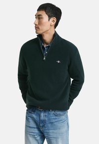 Man in een donkergroene trui met kwart rits over een blauw denim shirt en lichtblauwe spijkerbroek, staand met handen achter zijn rug, kijkend naar links.