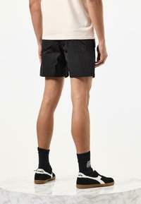 Schwarze Shorts aus leichtem Material mit strukturiertem Finish. Kombiniert mit schwarzen Sneakers mit weißen Akzenten und Gummisohle. Schwarze Socken sichtbar.
