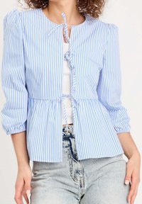 Blouse rayée bleu clair et blanc avec manches bouffantes, boutonnée à l'avant, et fermeture par lien. Confectionnée en tissu léger, avec un design à basque.