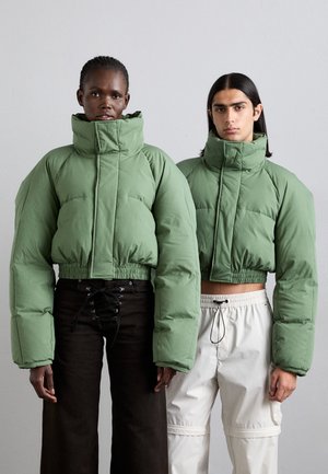 Jachete puffer verzi, scurte, cu guler înalt și tivuri elastice. Un model poartă pantaloni negri cu șireturi, iar celălalt pantaloni cargo de culoare deschisă.