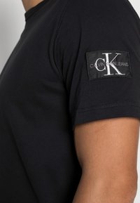 Černé tričko s krátkým rukávem vyrobené z bavlny, s látkovou nášivkou s logem Calvin Klein Jeans na levém rukávu. Hladká textura.