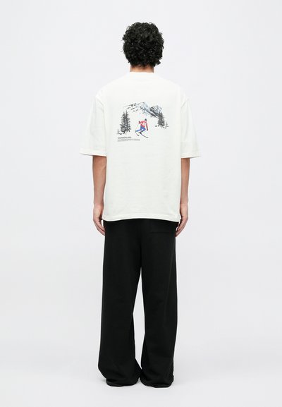 Svetlá oversized tričko s grafickou potlačou lyžiara, obsahujúcou prvky hory a stromov, v kombinácii s voľnými čiernymi nohavicami.