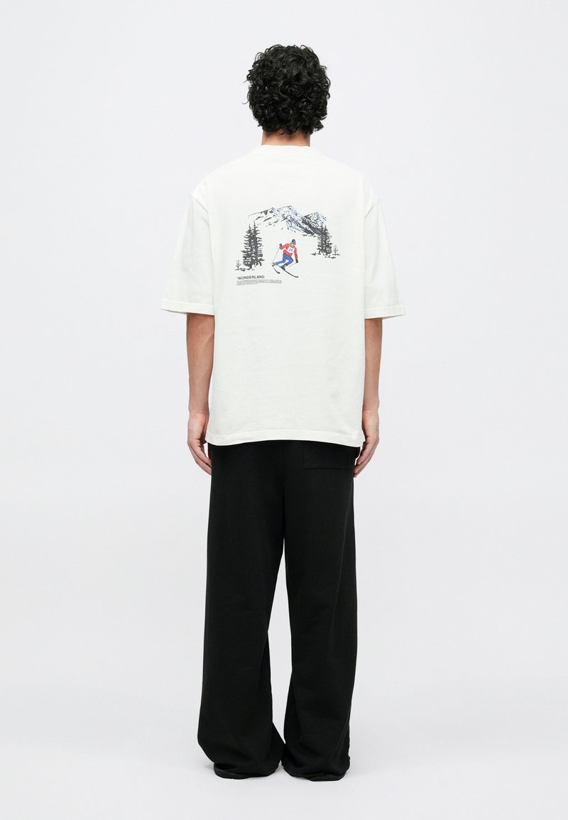 Maglietta oversize di colore chiaro con stampa grafica di uno sciatore, con elementi di montagna e alberi, abbinata a pantaloni larghi neri.