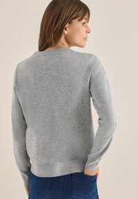 Donna di profilo che indossa un maglione a maniche lunghe grigio chiaro e jeans blu, con una mano nella tasca posteriore.