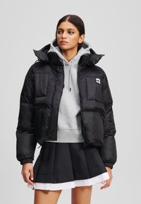 Doudoune noire avec un col haut, silhouette surdimensionnée et poches zippées. Portée par-dessus un hoodie gris et une jupe plissée noire.