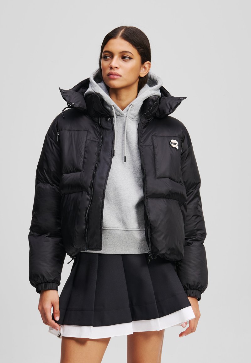 Doudoune noire avec un col haut, silhouette surdimensionnée et poches zippées. Portée par-dessus un hoodie gris et une jupe plissée noire.