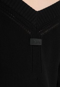 Svart stickad tröja med djup V-ringning, ribbad halslinning och en vävd etikett sydd i tyget. Texturerat mönster genom hela.