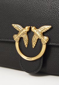 Black textured leather bag featuring a gold circular clasp with two intricately designed birds. Brand label "PINKO" visible on clasp.