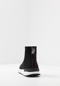 Black ankle-height sneaker with a textured knit upper, contrasting red stitching, and a white midsole with a black outsole.