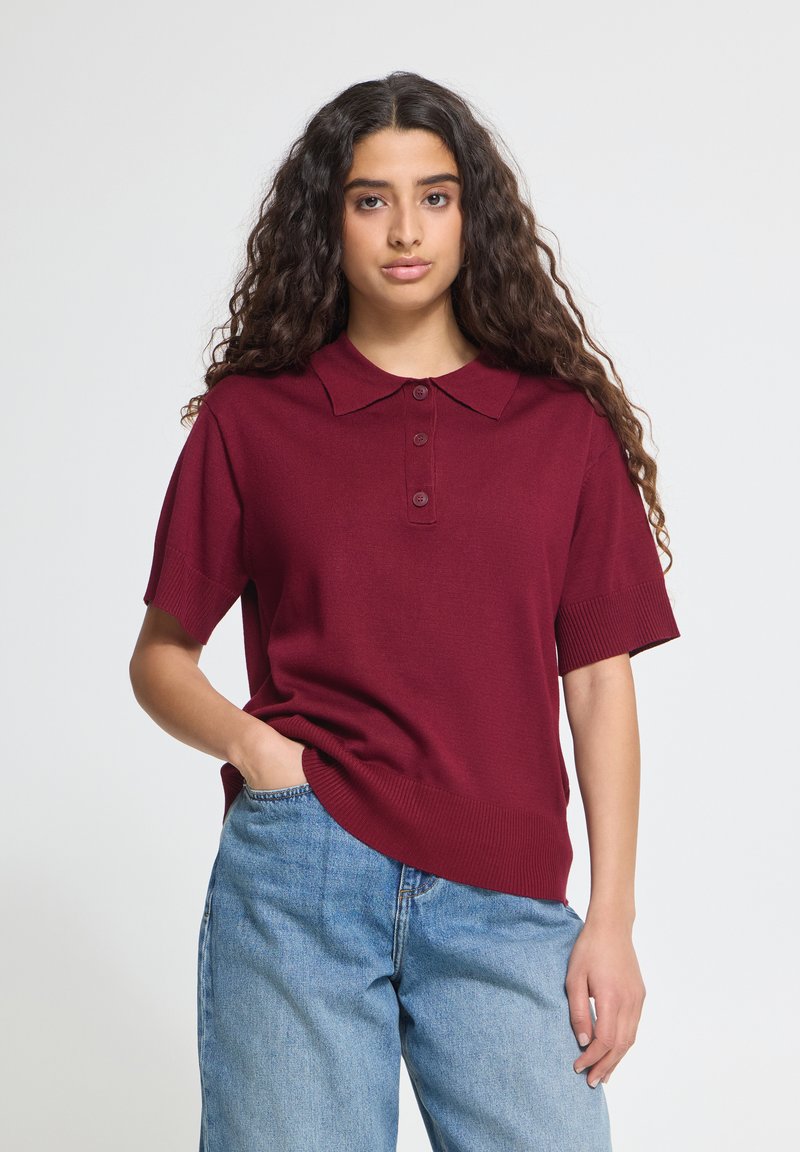 Femme aux longs cheveux bouclés foncés, portant un polo bordeaux à manches courtes et un jean bleu clair, debout avec une main dans la poche.
