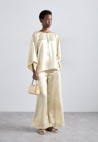 Cream satin top with wide sleeves and a keyhole neck; matching wide-leg pants; accessorized with a small beige handbag and strappy sandals.