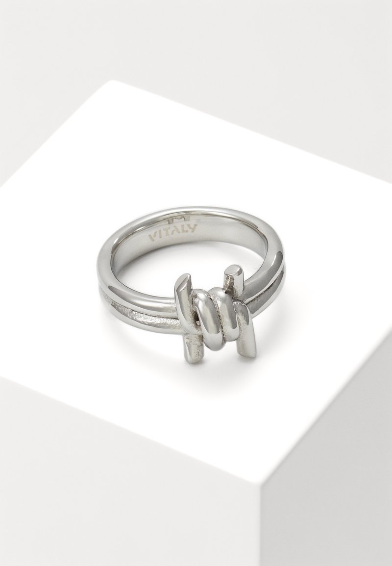 Bague en argent avec un détail de conception en fil barbelé sur une bande lisse, présentée sur un support géométrique blanc.